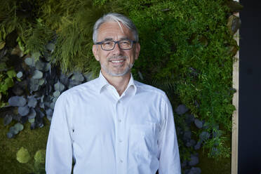 Happy mature businessman standing in front of plant wall at workplace - RBF08450