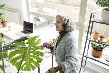 Senior businesswoman with headphones spraying water on plants at home - JCCMF04761