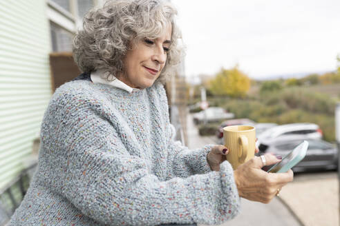 Ältere Frau mit Kaffeebecher und Smartphone auf dem Balkon zu Hause - JCCMF04782