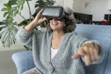 Senior woman wearing VR glasses gesturing on sofa at home - JCCMF04791