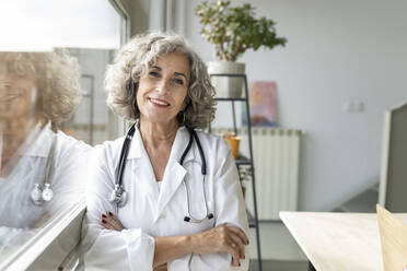 Smiling senior doctor with arms crossed leaning by window in home office - JCCMF04794