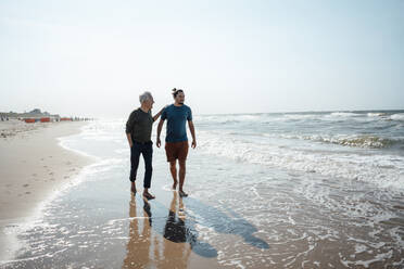 Father and son walking together on wet sand at beach - GUSF06563