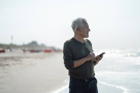 Älterer Mann mit Smartphone am Strand stehend - GUSF06566