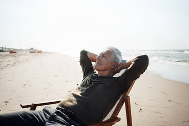 Senior man with hands behind head relaxing on chair at beach - GUSF06575