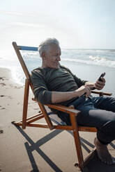 Älterer Mann mit Smartphone auf einem Stuhl am Strand sitzend - GUSF06579