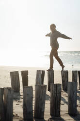 Junge Frau mit ausgestreckten Armen, die auf Holzpfählen am Strand läuft - GUSF06581