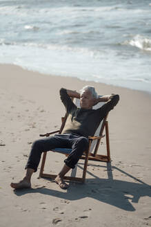 Älterer Mann mit Händen hinter dem Kopf auf einem Stuhl am Strand - GUSF06594