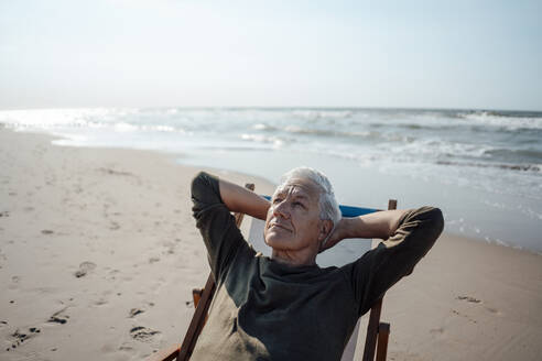 Älterer Mann blickt auf und entspannt sich auf einem Stuhl am Strand - GUSF06596