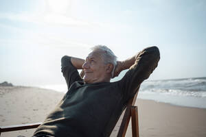 Lächelnder älterer Mann mit Händen hinter dem Kopf beim Entspannen am Strand - GUSF06597