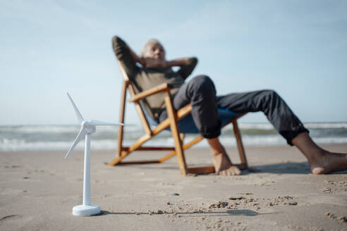 Windradmodell auf Sand mit einem älteren Mann, der sich am Strand entspannt - GUSF06598