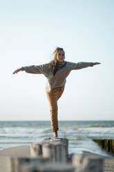 Woman with arms outstretched balancing on wooden post at beach - GUSF06628