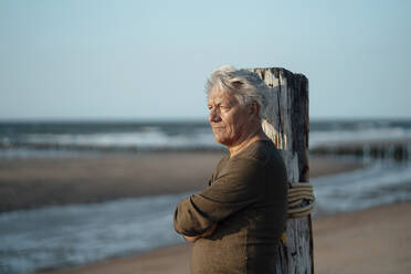 Älterer Mann lehnt an einem Holzpfosten am Strand - GUSF06632