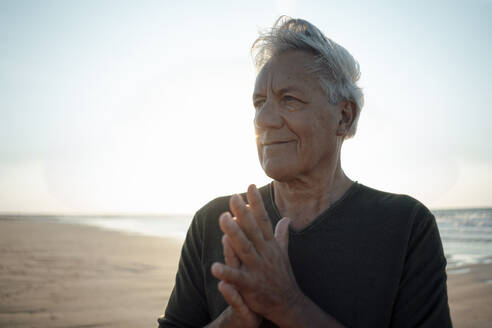 Smiling senior man with hands clasped at beach - GUSF06636