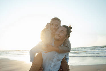 Smiling man piggybacking woman at beach - GUSF06650