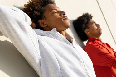 Seitenansicht von entspannenden jungen afroamerikanischen Freunden, die die Augen schließen, während sie gemeinsam an einer weißen Wand auf einer Straße im Sonnenschein schlafen - ADSF32538