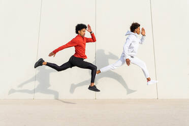 Side view of black male athletes in sportswear jumping against white wall during jogging in city - ADSF32540