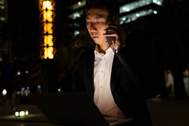 Fokussierter asiatischer männlicher Unternehmer in formeller Kleidung, der auf einem Laptop surft und telefoniert, während er auf einer Straße mit leuchtenden Gebäuden in der Nacht sitzt - ADSF32541