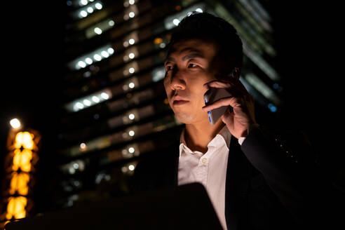 Fokussierter asiatischer männlicher Unternehmer in formeller Kleidung, der auf einem Laptop surft und telefoniert, während er auf einer Straße mit leuchtenden Gebäuden in der Nacht sitzt - ADSF32543