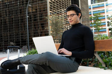 Seriöser asiatischer männlicher Unternehmer, der auf einem Netbook tippt, während er an der Grenze in der Nähe eines Koffers auf einer Straße mit modernen Gebäuden in der Stadt sitzt - ADSF32548