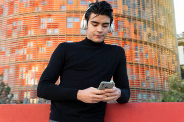 Gut aussehender asiatischer Mann, der mit drahtlosen Kopfhörern Musik hört, während er mit seinem Handy auf der Straße in der Nähe eines modernen Gebäudes in der Stadt Nachrichten sendet - ADSF32550