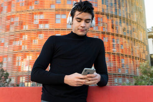 Gut aussehender asiatischer Mann, der mit drahtlosen Kopfhörern Musik hört, während er mit seinem Handy auf der Straße in der Nähe eines modernen Gebäudes in der Stadt Nachrichten sendet - ADSF32550