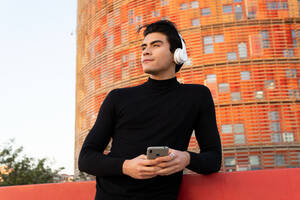 Gut aussehender asiatischer Mann, der mit drahtlosen Kopfhörern Musik hört, während er mit seinem Handy auf der Straße in der Nähe eines modernen Gebäudes in der Stadt Nachrichten sendet - ADSF32551