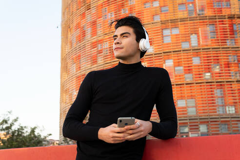 Handsome Asian male listening to music in wireless headphones while ext messaging on cellphone on street near modern building in city - ADSF32551