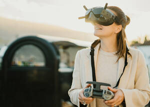 Positive anonyme Frau in modernem schwarzen VR-Headset mit Fernbedienung, die auf der Straße einer Stadt steht, während sie die virtuelle Realität erkundet - ADSF32556