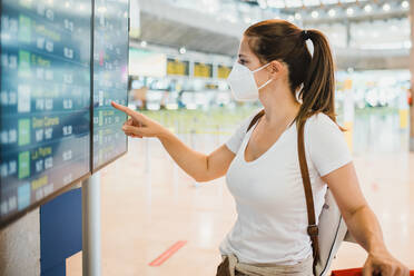 Side view of female traveler in protective mask standing near signboard with flights schedule in airport terminal before departure - ADSF32557