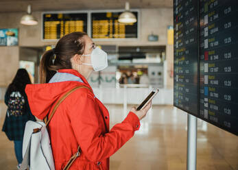 Seitenansicht einer weiblichen Reisenden mit Schutzmaske, die mit ihrem Smartphone in der Nähe eines Schildes mit dem Flugplan im Flughafenterminal vor dem Abflug steht - ADSF32558