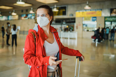 Side view of focused female in protective mask surfing cellphone while standing in modern airport terminal with suitcase during coronavirus pandemic - ADSF32559