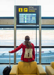 Rückenansicht einer anonymen weiblichen Touristin mit Rucksack und Koffer, die auf einem Sessel in der Nähe eines Schildes mit einem Flugplan im Flughafen sitzt - ADSF32563