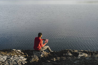 Mann liest ein Buch, während er auf einem Felsen am Seeufer sitzt - MASF27213