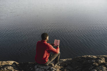 Rear view of man reading book while sitting on rock at lakeshore - MASF27214