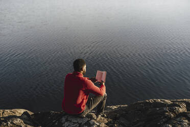 Rückansicht eines Mannes, der auf einem Felsen am Seeufer sitzt und ein Buch liest - MASF27214