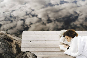 Hoher Blickwinkel der Frau, die ein Buch liest, während sie auf einer Wolke liegt, die sich auf einem See spiegelt - MASF27240