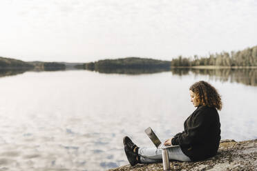 Frau arbeitet am Wochenende ferngesteuert am Seeufer an einem Laptop - MASF27253
