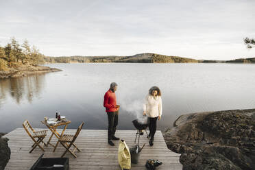 Full length of couple preparing food on barbecue grill against lake - MASF27267