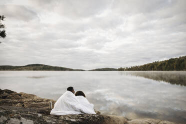 In eine Decke eingewickeltes Paar sitzt am Seeufer vor bewölktem Himmel - MASF27275