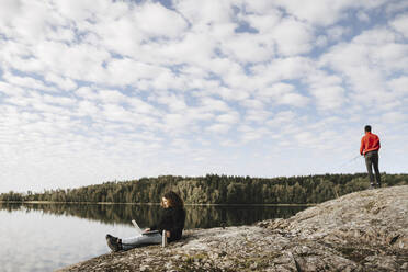 Woman working remotely on laptop while boyfriend fishing at lakeshore - MASF27294