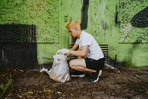 Seitenansicht eines jungen männlichen Freiwilligen in der Hocke beim Aufsammeln von Plastikmüll an einer grünen Wand - MASF27360