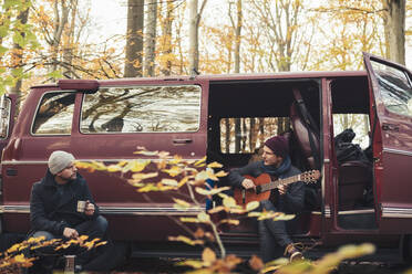 Mann spielt Gitarre, während sein Freund beim Camping im Wald etwas trinkt - MASF27423