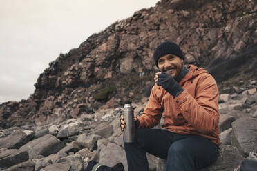 Smiling man having drink while sitting on rocks at beach - MASF27447