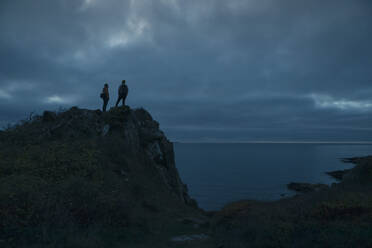 Mature male friends standing on top of hill by sea at dusk - MASF27454