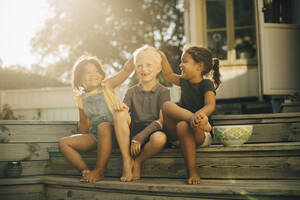Playful sisters with male friend sitting on steps at patio during sunny day - MASF27495