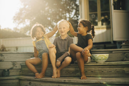 Playful Schwestern mit männlichen Freund sitzen auf Stufen auf der Terrasse während sonnigen Tag - MASF27495