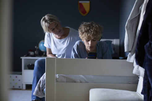 Mother looking at son using smart phone on bed in bedroom - MASF27545