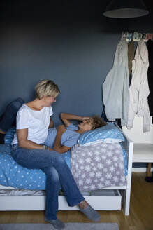 Smiling mother talking to son lying on bed at home - MASF27549