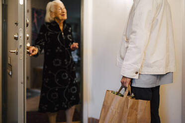 Ältere Frau, die die Tür öffnet, während ein Pfleger mit Lebensmitteln zu Hause ankommt - MASF27637