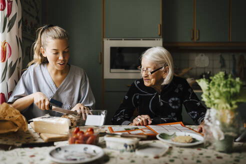 Weibliche Pflegekraft schneidet Brotlaib am Esstisch, während sie neben einer älteren Frau in der Küche sitzt - MASF27642